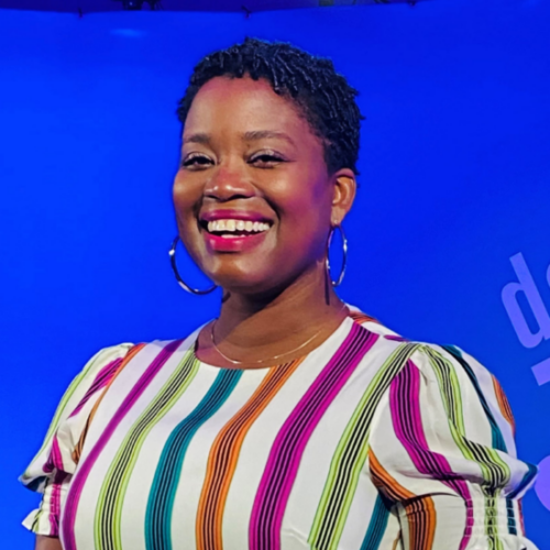 Headshot of a smiling person of dark skin tone with short black hair in braids, wearing hoop earrings, red lipstick, and a striped shirt in vibrant shades of blue, pink, orange and green.