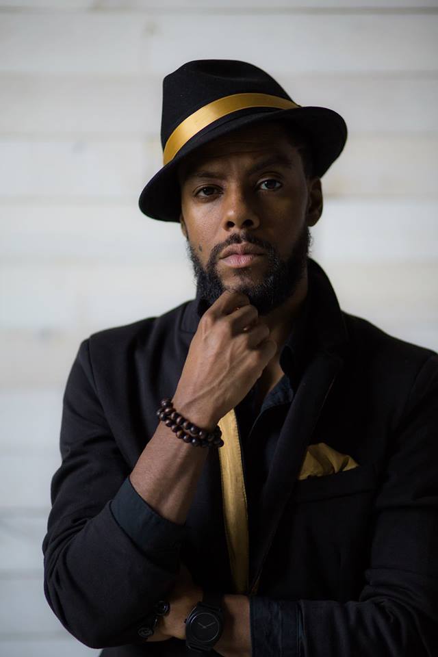 Headshot of a person of dark skin tone and a short black mustache/beard, with a pensive look on their face and hand raised to chin, wearing a black fedora like hat and suit with gold accents.