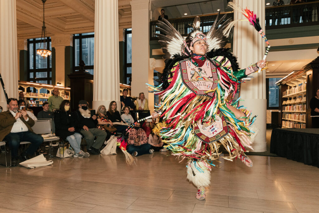 A dancer in full Native Amerian regalia performs for a crowd in a packed library.