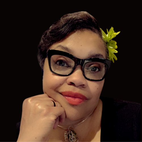 Headshot of a person with a soft smile, of medium light skin tone and short dark hair, wearing a green flower clip in their hair, as well as large dark frame glasses, light red lipstick, and a necklace.