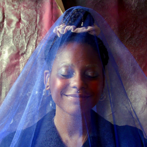 Headshot of a smiling person with their eyes closed, of dark skin tone and black braided hair up in a bun, wearing a blue veil over their head, silver earrings, a piercing above their lip, and a black top.
