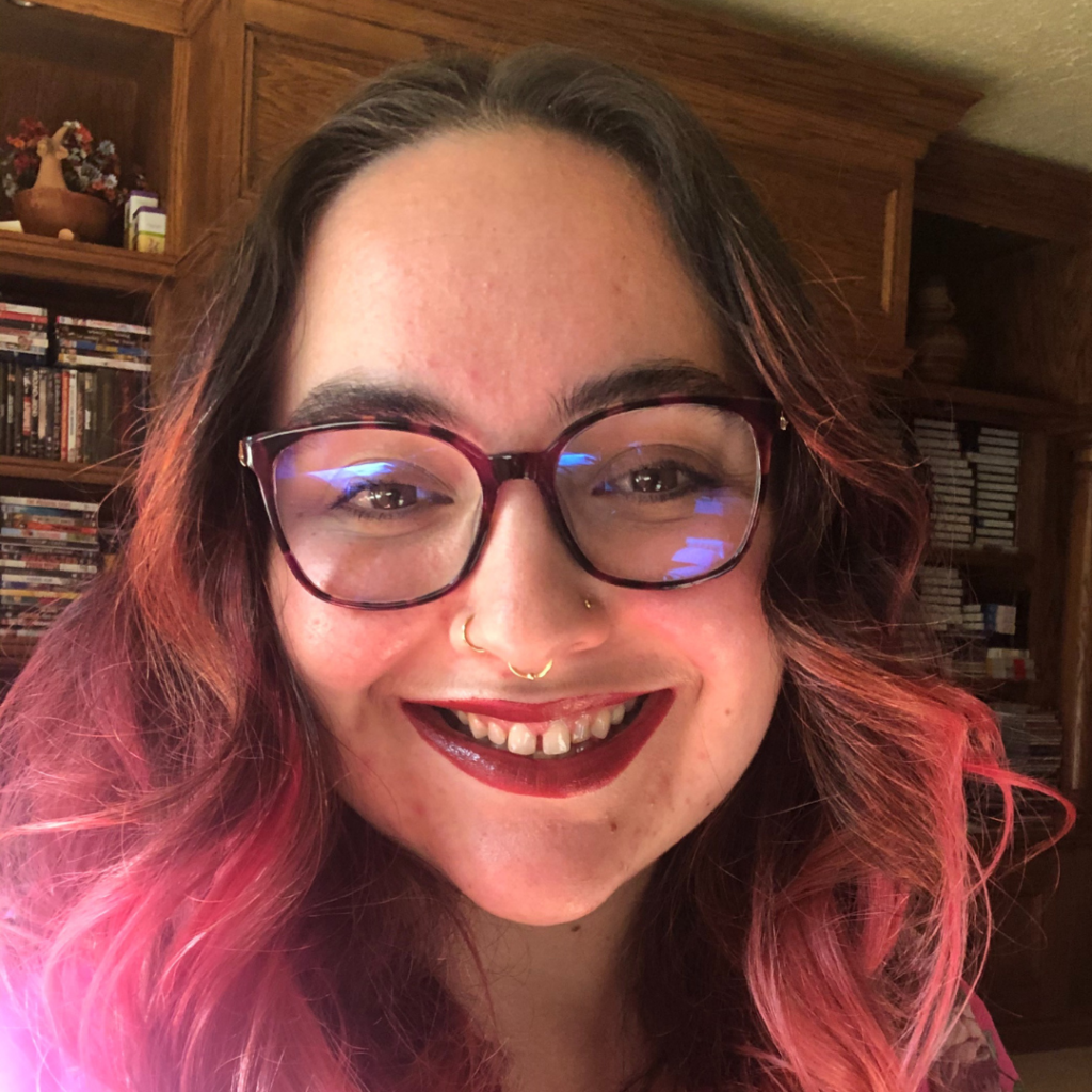 Headshot of a smiling person of light skin tone and long wavy pink hair, with maroon glasses and nose piercings, wearing red lipstick and standing in front of a bookshelf.