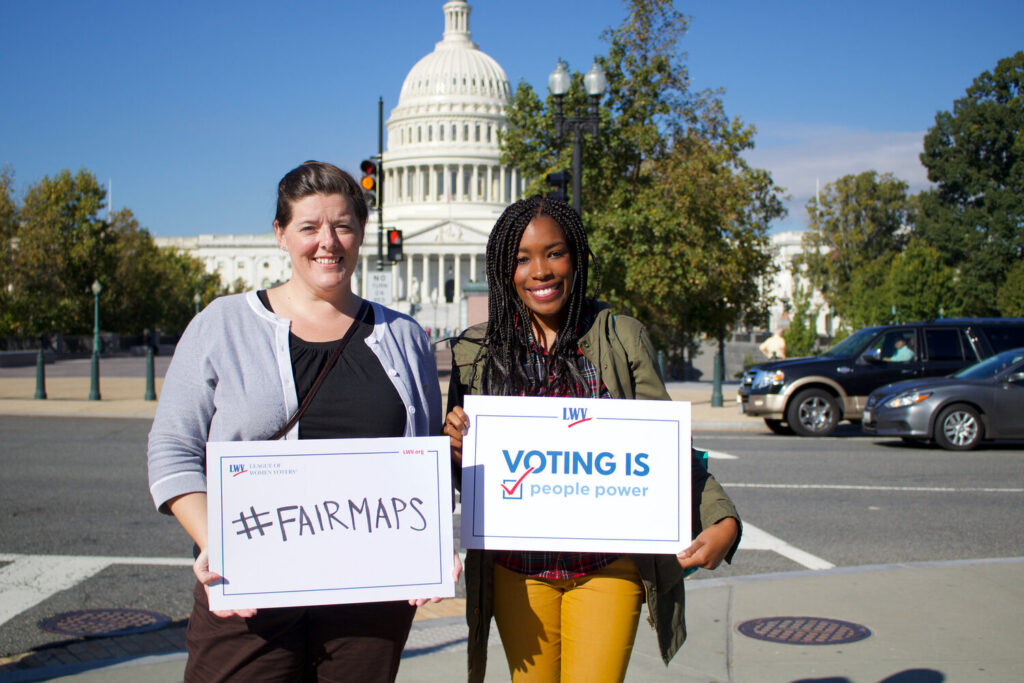 Two people from League of Women Voters US stand with signs that read "#FairMaps," and "Voting is People Power." The Minnesota state capitol building can be seen in the background