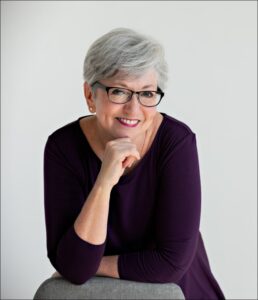 Headshot of a smiling person of light skin tone and short white hair, with dark framed glasses and wearing a dark purple dress, leaning on a chair with the one hand up to their chin.