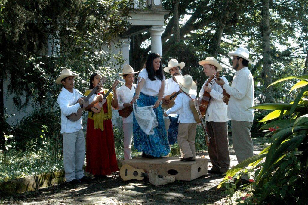Los Utrera, a band based in Mexico whose music and dance is part of the son jarocho tradition—a multicultural mix of Spanish, African, and indigenous influences, perform outside amongst trees and plants. Two people are dancing on a wooden platform while others perform on guitar and various other instruments around them.