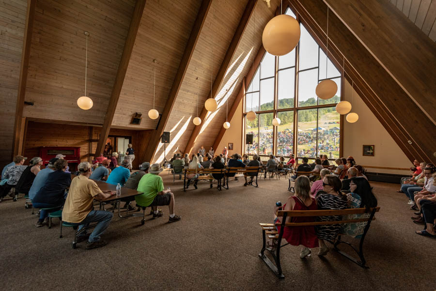 Many people sit at tables and benches inside of a retreat center, facing towards people sitting on a stage and speaking the corner.