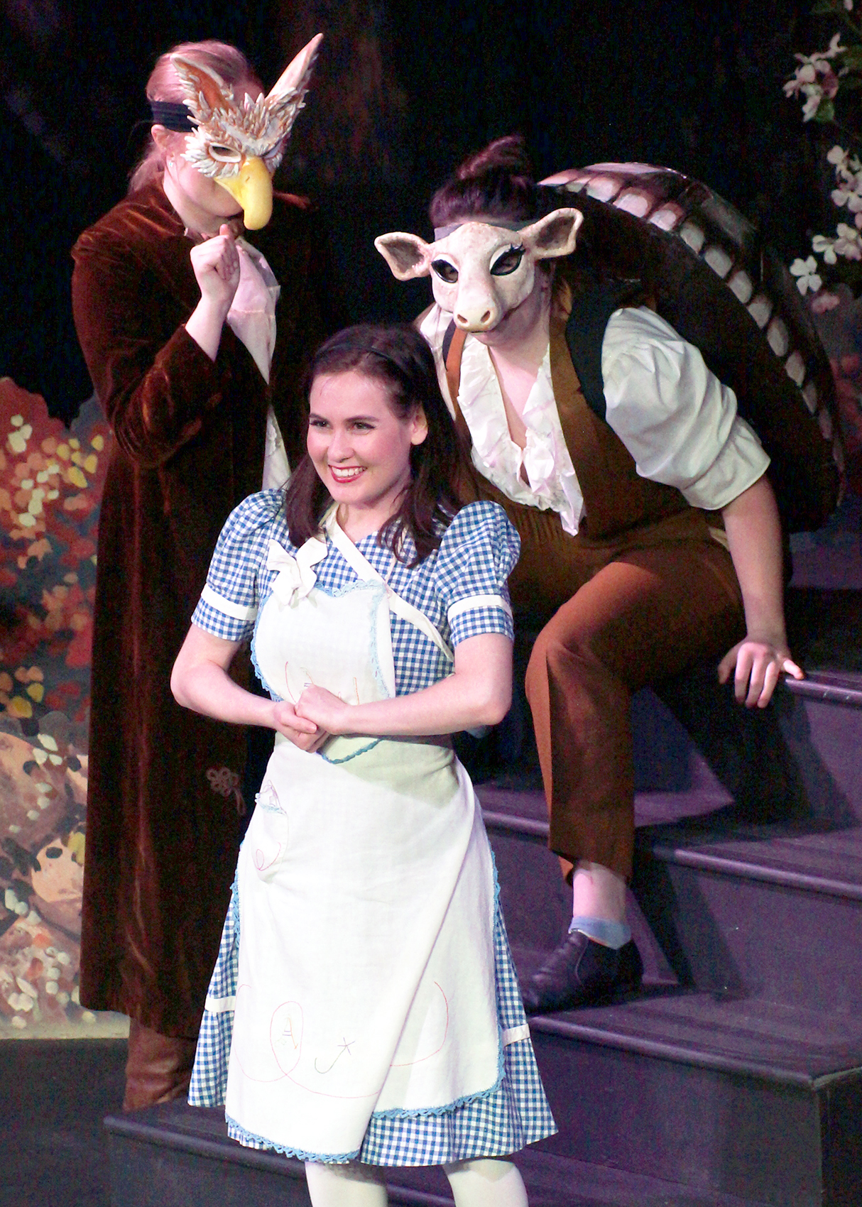 Performance of "Alice in Wonderland" at the Matthews Opera House in Spearfish, South Dakota. One smiling person wearing an apron over a blue checkered dress stands with hands held together. Behind them there are two people leaning in and wearing animal masks.