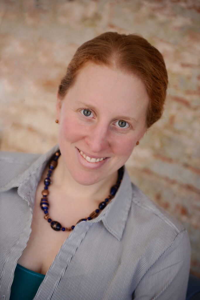 Headshot of a smiling person of light skin tone with red hair pulled back, wearing a gray collared shirt and a beaded necklace.