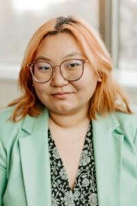 A smiling person of medium light skin tone, with light brown rimmed glasses and shoulder length pink hair, wearing a seafoam green blazer over a black and seafoam green patterned top