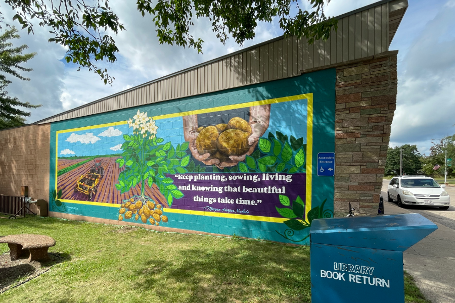 Rectangular-shaped mural on the back of a brick building. In the center, there’s a pile of seeds with leaves and flowers growing from them. To the left there is a scene of a tractor driving through a farm field under a blue and cloudy sky. To the right there are two hands holding a pile of potatoes. Below that are the words, “‘Keep planting, sowing, living and knowing that beautiful things take time.’-Morgan Harpor Nichols.” *Part of the Women’s Suffrage Centennial Commission*
