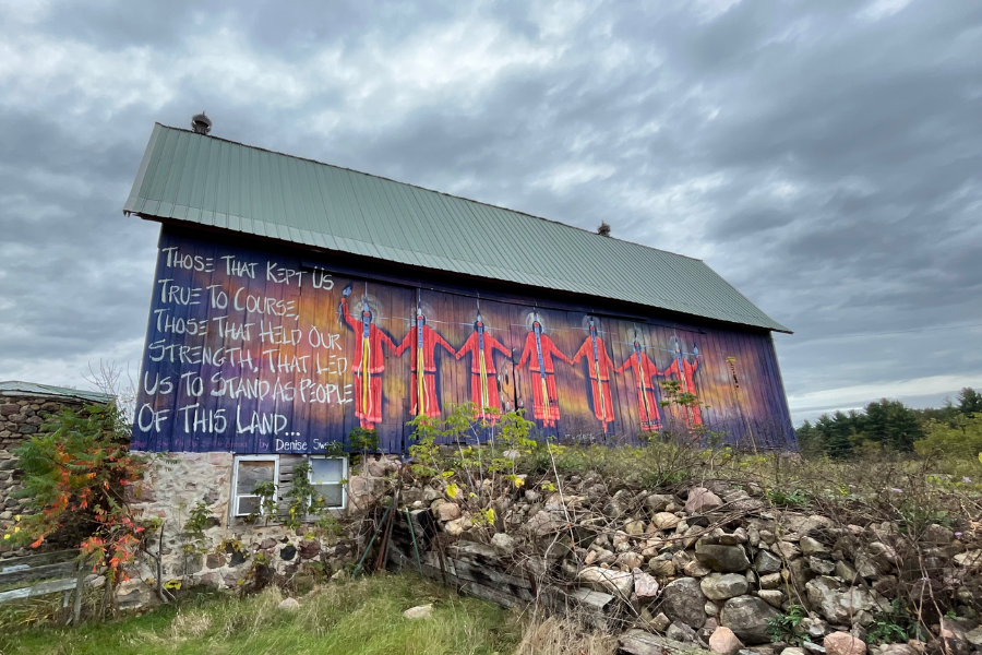 Mural on the side of a farmhouse surrounded by plants and rocks. The background is a dark purple, with an orange glow surrounding seven people who stand hand in hand. Each of them wear the same red outfit and have a white glow around their heads. To their left, in white paint, are the words “Those that kept us true to course, those that held our strength, that led us to stand as people of this land…by Denise Sweet” *Part of the Women’s Suffrage Centennial Commission*