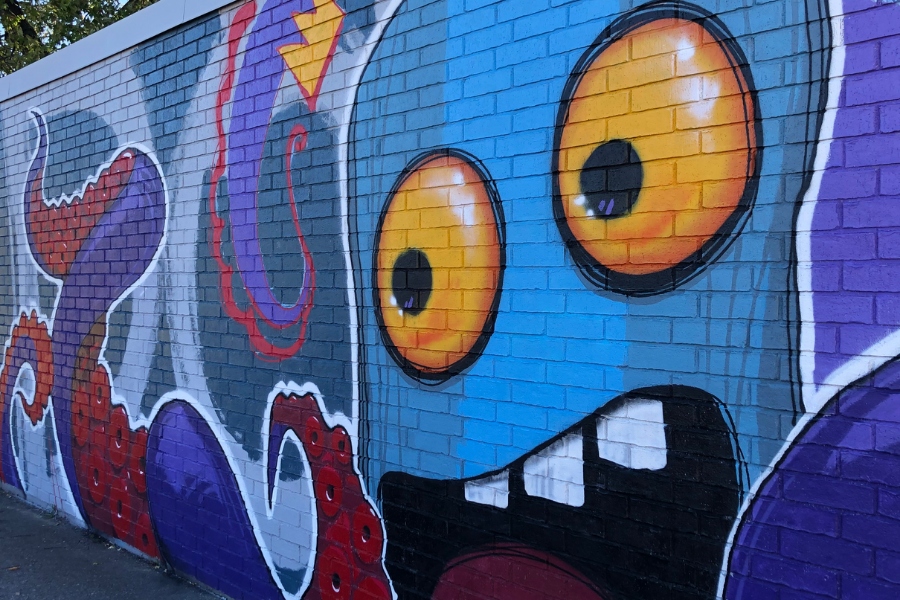 Mural of a close up of a blue octopus on a brick wall. The octopus’ head is light blue and it has big circular orange eyes. Its mouth is wide open, showing three teeth, making it look shocked or scary. There’s a small crown floating above its head to the right. Its tentacles are twisting around it and are dark blue with red suction cups.