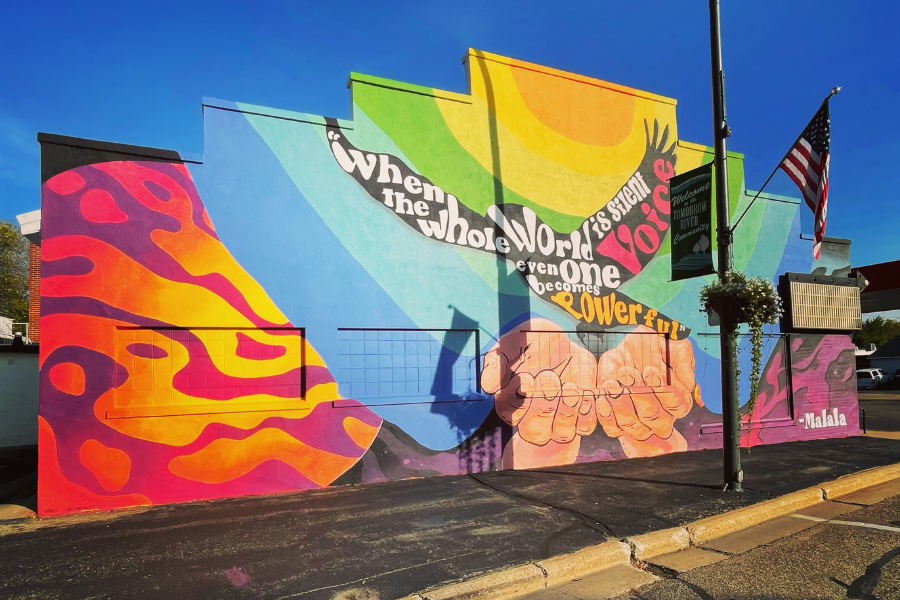 At the center of the mural are two open hands, palms facing up. It looks like the hands are freeing a bird. The bird is painted black with the words “When the whole world is silent even one voice becomes powerful” over it in white, pink and orange paint. The background is striped with rainbow colors and a pattern of orange and purple paint that resembles a butterfly wing. *Part of the Women’s Suffrage Centennial Commission*