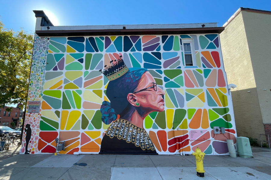 Mural on the side of a building showing a profile view of Ruth Bader Ginsburg at the center. Different colors swirl and contour her face and hair, which is up in a bun. She has a stoic look on her face. She’s wearing a crown and the spikes are replaced with miniature silhouettes of people protesting with signs. She’s wearing a decorative piece around her neck which says “Women belong in all places where decisions are being made. – RBG” Vibrant, multi-colored shapes are grouped in squares around her and cover the rest of the mural. *Part of the Women’s Suffrage Centennial Commission*