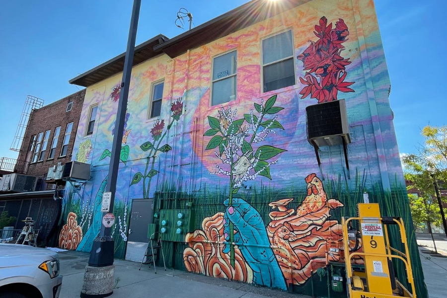 Multi-colored mural on the back of a tall building. Amongst tall grass at the bottom is a large blue colored hand with pink nails. The hand is holding a leafy plant of some kind, and two goat heads are depicted sprouting from the plant. Behind the hand there’s dark orange swirly fungi with two chickens seemingly growing from it. Above, there’s a red flowery plant with three heads of cardinals poking out. To the left, there’s another plant with leaves that have eyeballs. The sky is a swirling cloudy sunset of blues, pinks, purples, and yellows.