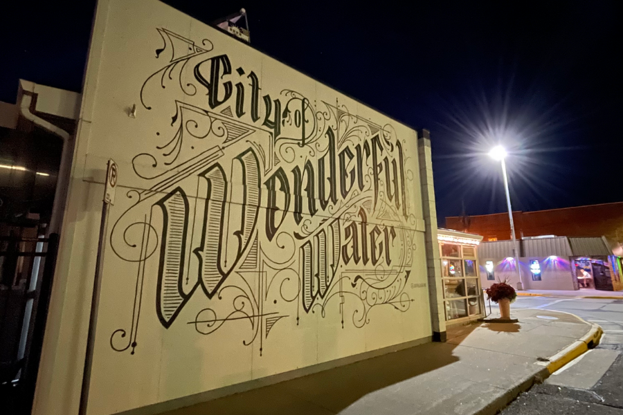 Mural on the side of a small building at nighttime. The background is a cream white, and written in large, black, elaborate font are the words “City of Wonderful Water.”