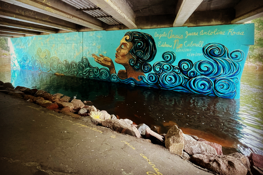Mural under an overpass bridge in a river. It is painted light blue, with a woman of medium skin tone and black curly hair at the center. She’s blowing dandelion seeds off of her hand, which float off into the distance. It looks like she’s in water, as swirly designs resembling waves, and closely matching the pattern of her hair, come up to meet her shoulders. To her right, in yellow paint, are the words “Angela Anaïs Juana Antolina Rosa Edelmira Nin Culmel” and the dates “2/21/1903-1/14/1977.” *Part of the Women’s Suffrage Centennial Commission*