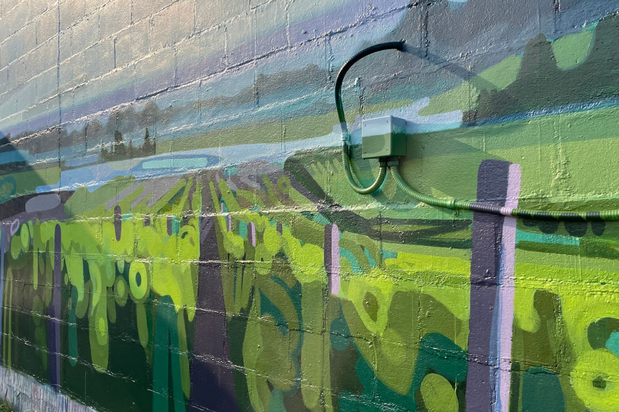 A close up of a mural on a brick building. The painting is of a farm field; rows of bright green stalks lead up to blue body of water. There are hills in the distance that blend into the blue and purple sky.