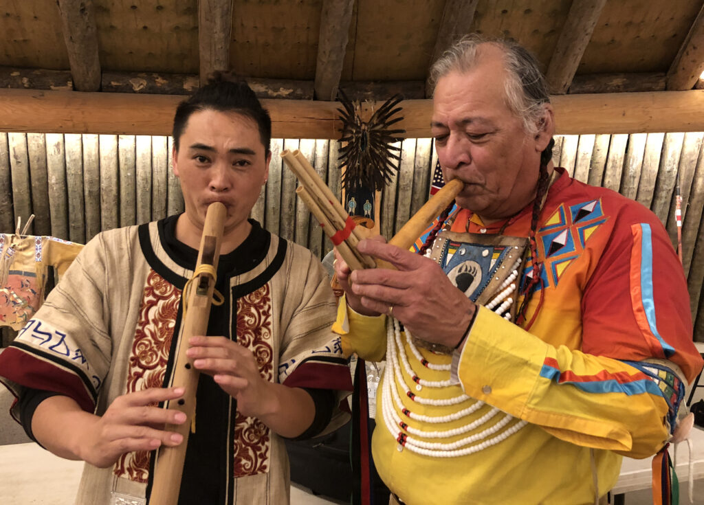 Two people stand next to each other and play different types of wooden flutes.