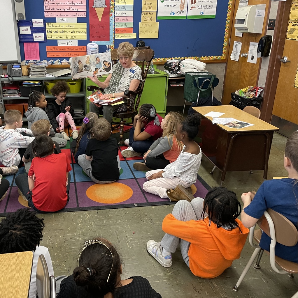 An adult is sitting in a rocking chair in a classroom, reading a book to a group of young children seated on the floor.