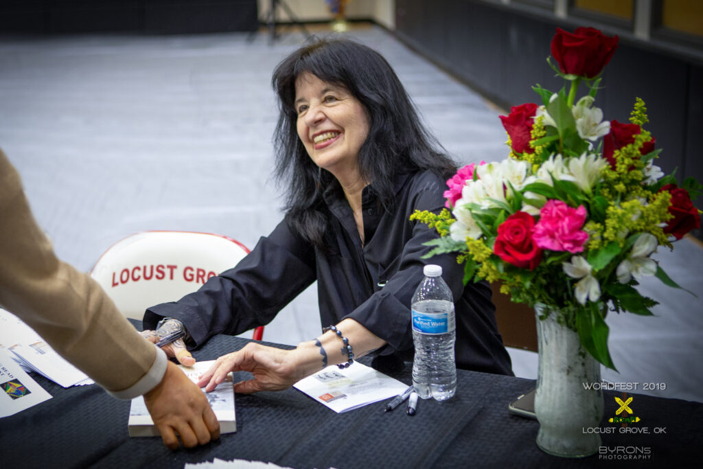 A person sits at a table next to a bouquet of flowers, smiling up at a person whose book they're about to sign.
