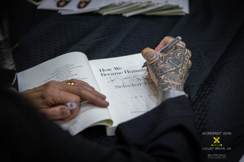 Close up of a tattooed hand signing the inside page of "How we Became Human" by Joy Harjo.