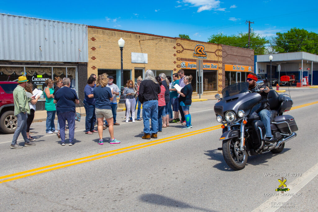 A person on a motorcycle drives past a group of people gathered together in the street, blocking a car lane outside of a consignment shop, and holding up books in their hands.