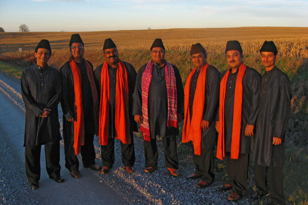Qawal Najmuddin Saifuddin & Bros, a qawwali ensemble from Pakistan, stand together in front of a meadow and pose.
