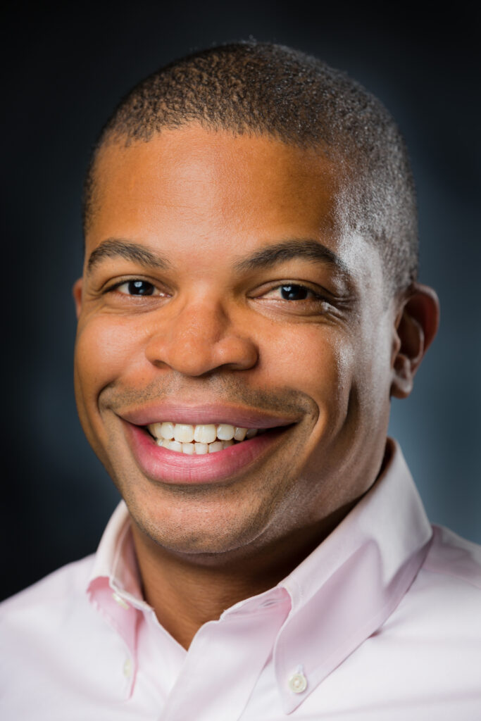Headshot of a smiling person of medium dark skin tone and buzzed dark hair, wearing a light pink button down shirt.