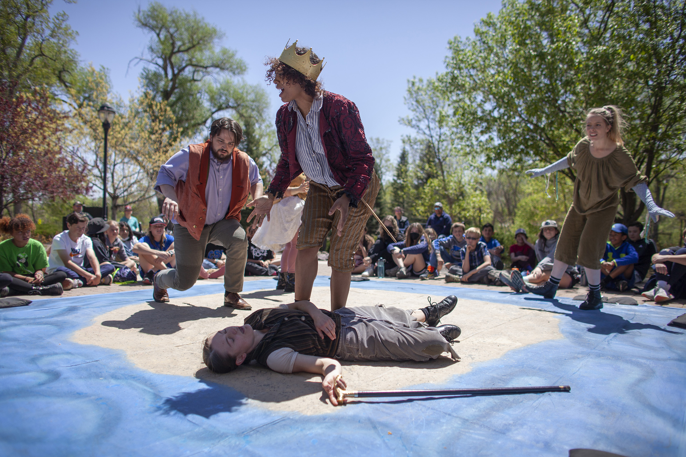 People perform on a cement square outside with students sitting around them. In the center there's a person wearing a crown and Shakespeare era clothing, seemingly crying out as a person with a cane lays at their feet, appearing to be dead.