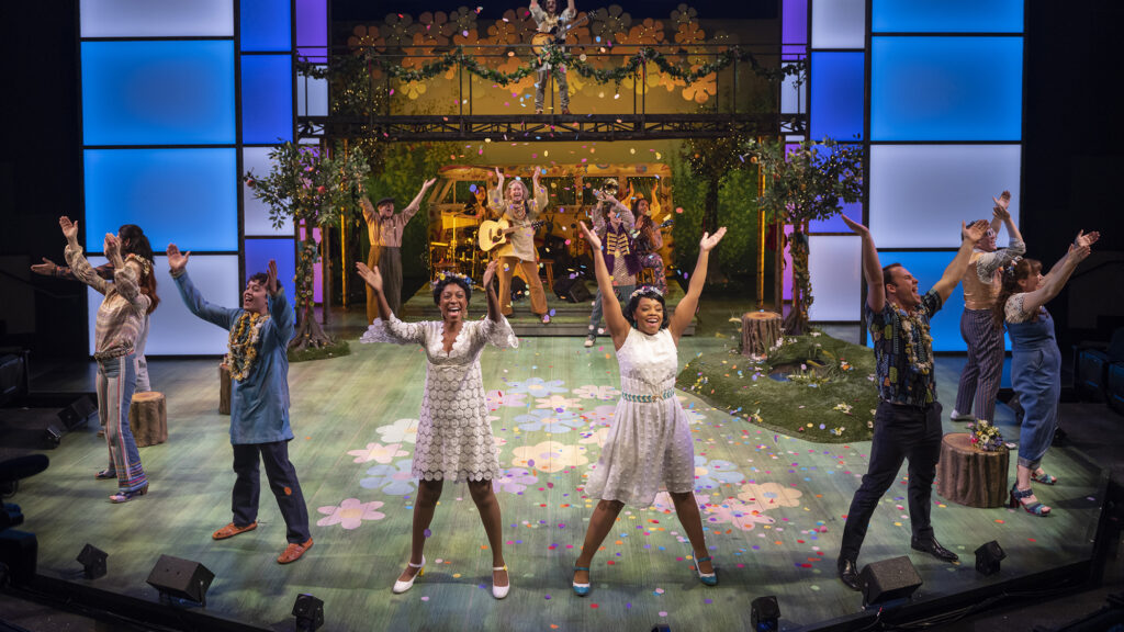 Eight people stand in a semicircle at the top of a stage, with their hands in the air and smiles on their faces. Behind them, there's a stage where people are performing with musical instruments and also have their hands in the air. The stage is set up with fake trees, grass, logs, and painted flowers, and there's confetti falling from the sky; all creating a Spring like vision.