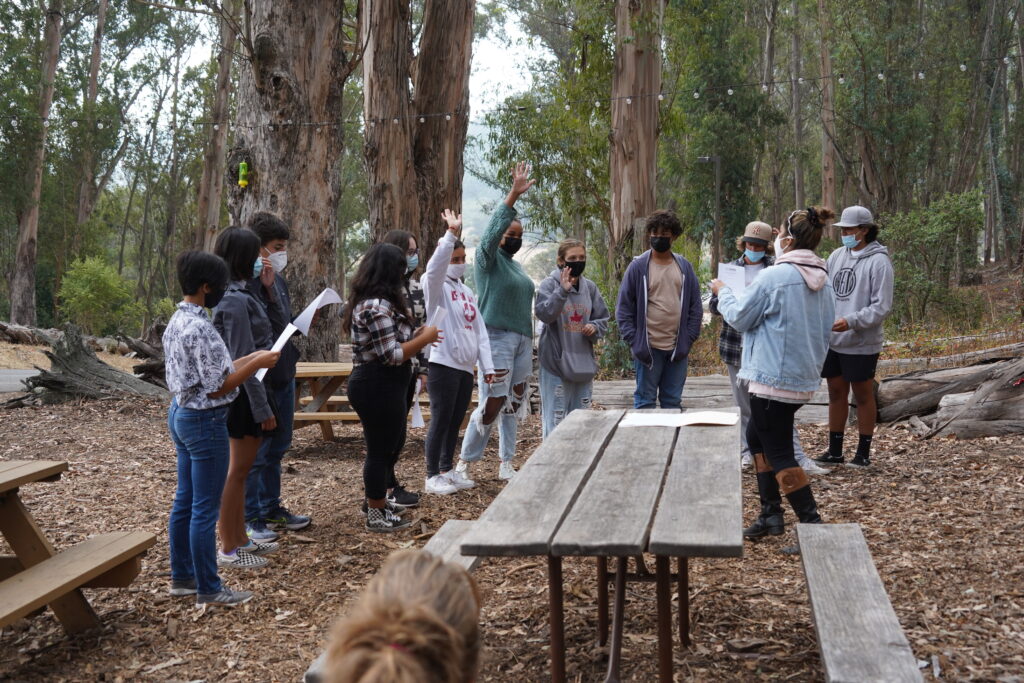 A group of people in protective masks stand together in a clearing in the woods. Some are raising their hands and others are looking at pieces of paper. There are picnic benches and twinkling lights hanging around them.
