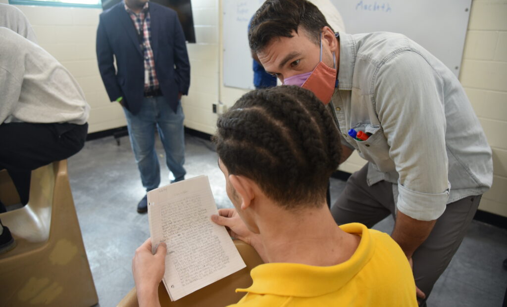In a classroom setting, a person wearing a protective cloth mask is leaning down and talking to a younger person who is holding sheets of loose leaf paper covered in writing.