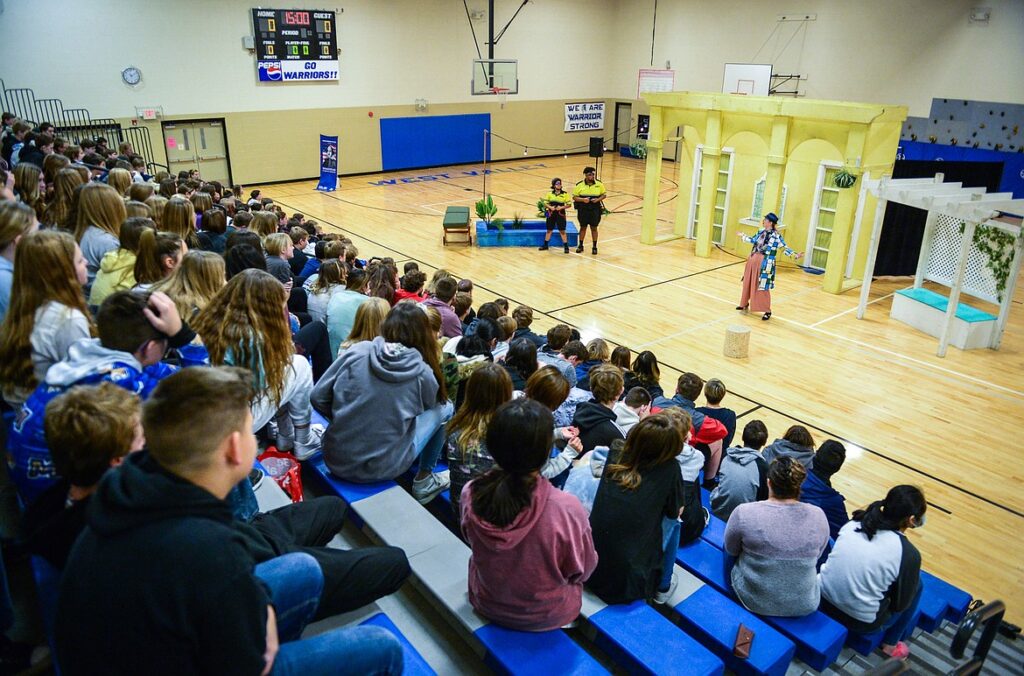 A crowd of students sitting on bleachers in a gymnasium watch a performance happening on the gym floor.