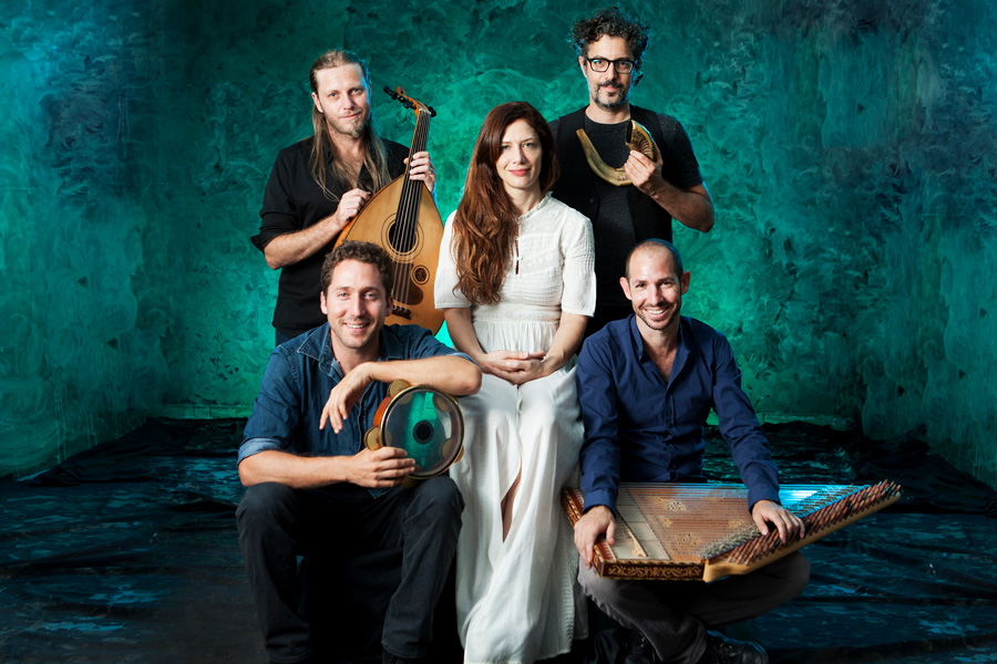 Sofi & the Baladis, a band based in Israel, pose together with various musical instruments in front of a green patterned wall.