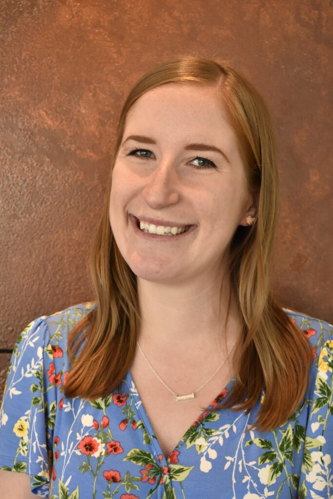 Headshot of a smiling person of light skin tone and shoulder length red hair, wearing a blue shirt with floral pattern and a necklace, standing in front of a brown wall.