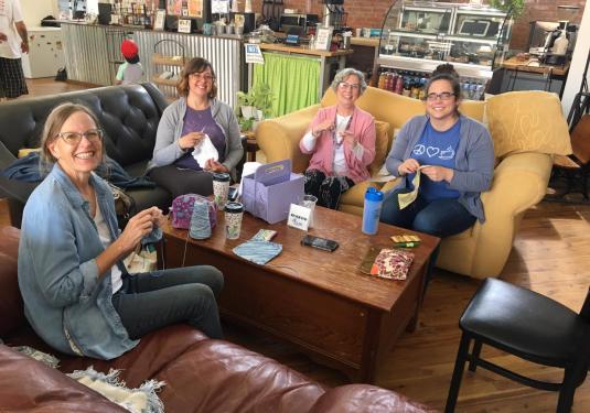 A group of four people sit on couches around a table in a coffee shop, holding up pieces that they're each stitching.