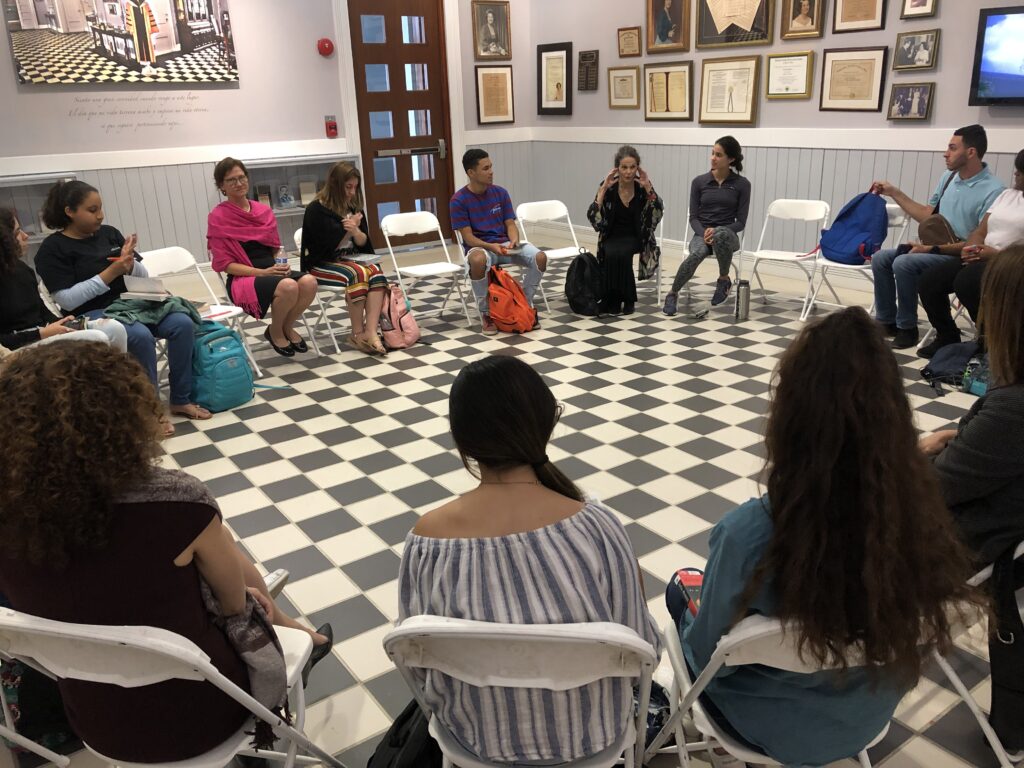 A group of students sit in chairs in a circle with Julia Alvarez, who is currently speaking.