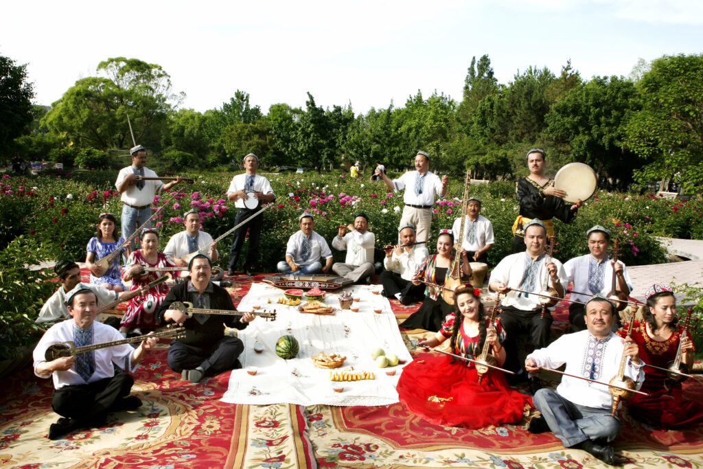 Tarim, a large band based in China, sit on rugs together in the middle of a garden with various fruits in the center, and pose with their instruments.