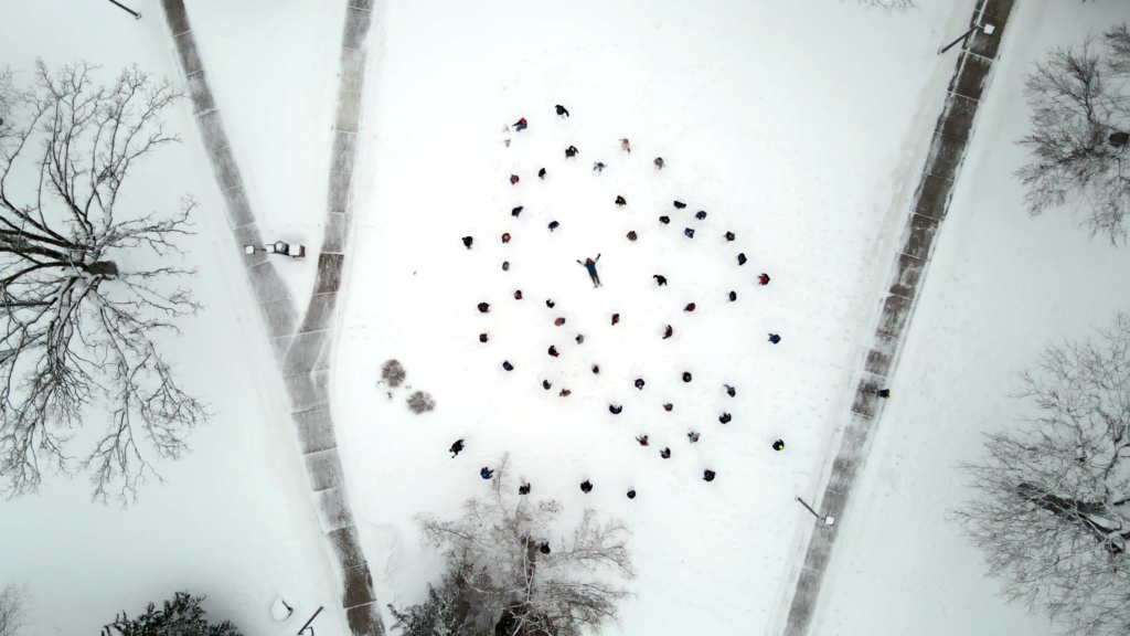 Snowy drone shot of UW-Stout Students from the Outro Carnaval Video. Students circle around one person in the center, who is laying on their back with arms and legs spread out, in snow angel like fashion.