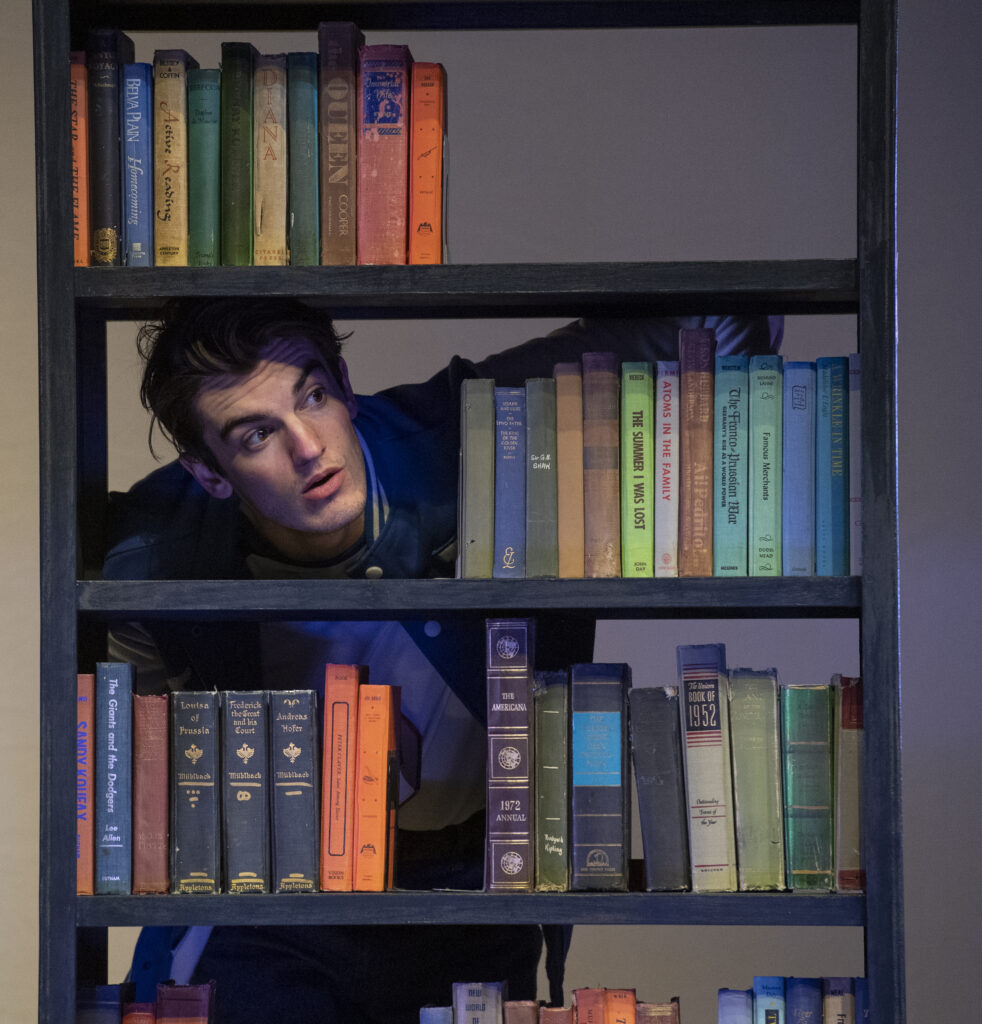A person stands behind a book shelf, with their face in an opening next to a stack of books, looking off to the side with a curious expression.
