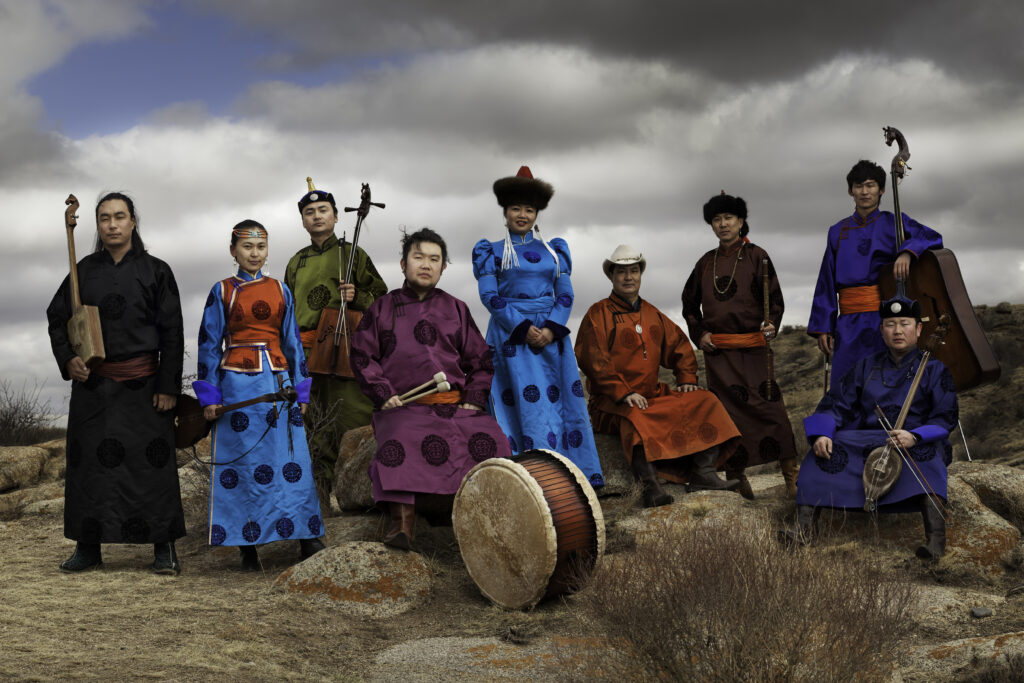 Anda Union, a band based in China who primarily specialize in khoomii (a traditional type of Mongolian overtone singing), pose in the middle of a mountain range, wearing vibrant shades of blue, purple, red, and green, holding various instruments.