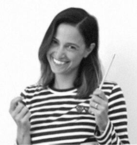 Headshot of a smiling person of light skin tone and shoulder length dark hair, with their hands up and one holding a pencil, wearing a black and white striped shirt.