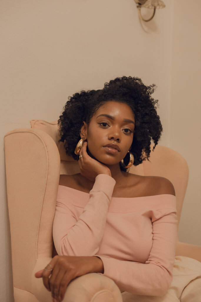 Headshot of a person with a gentle facial expression, of medium dark skin tone and short curly black hair, poses in a chair with one hand up to their face, wearing a light pink top that closely matches the chair.