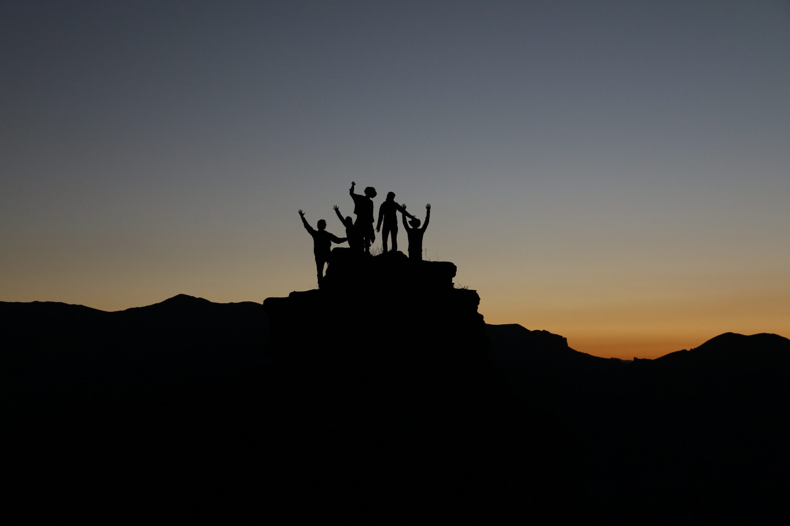 Silhouettes of five people standing on top of a rock formation at dusk, with their hands in the air.