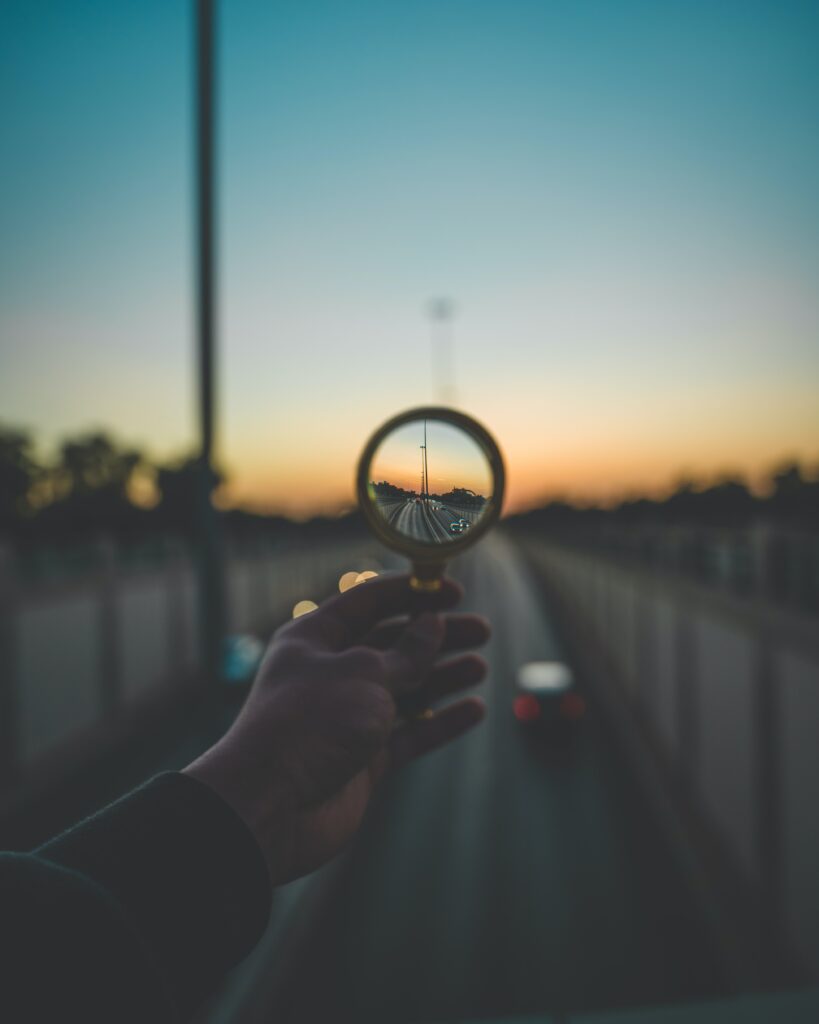 A hand holds a magnifying glass up over a highway at dusk. Everything is unfocused except for the view through the magnifying glass, which brings the distance into clarity.