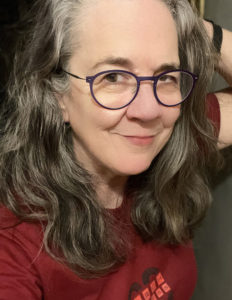 A white woman wearing glasses with dark greying hair looking into the camera with a soft smile.
