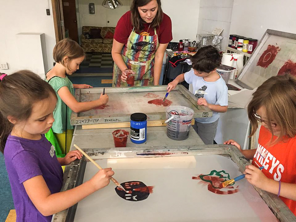 A group of four children paint on two silkscreens as they are guided by an adult.