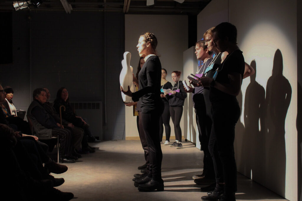 A group of theater performers, some holding white oblong vessels, stand in front of a seated audience.