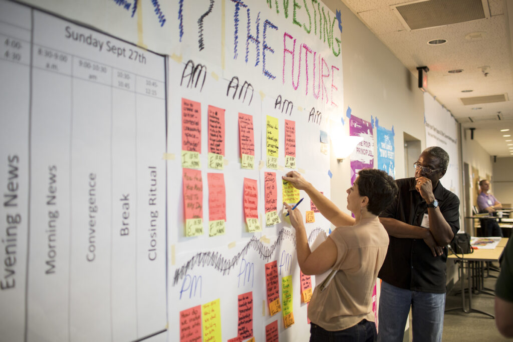 At the ArtsLab final retreat in September 2014, a person adds a post-it note with writing on it to a giant piece of poster paper, while a person behind them observes.
